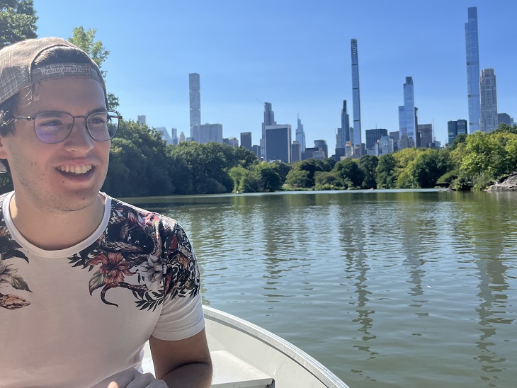 Fiatal férfi fehér pólóban és baseball sapkában egy tavon csónakázik New Yorkban háttérben a felhőkarcolókkal