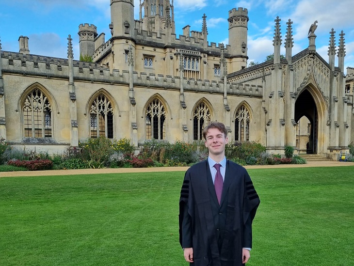 ösztöndíjas hallgató a Cambridgei Egyetem épülete előtt áll a zöld füvön