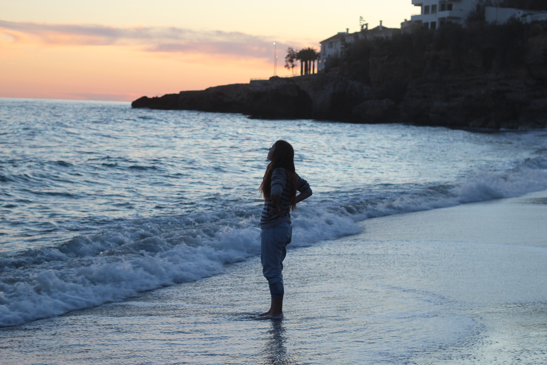 Vízparti naplemente a homokos parton álló lány lábát nyaldossák a hullámok háttérben egy kiszögellő részen fák és házak