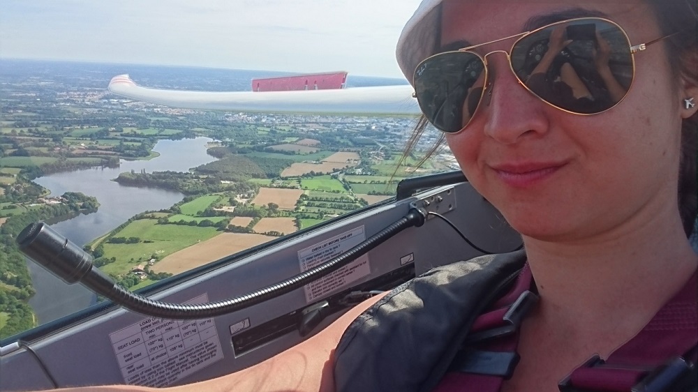 Fiatal lány repülőgépet vezetve szelfizik Alatta a váltakozó zöld mezős táj 